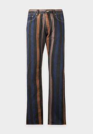 Jeune homme portant un t-shirt marron à manches courtes avec bordure blanche, un pantalon rayé multicolore et des chaussures noires avec des accents dorés, debout avec une main dans la poche.