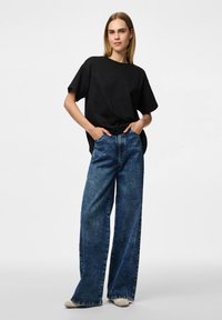 Zwart oversized t-shirt met korte mouwen, gecombineerd met hooggerande, wijde spijkerbroeken in denim blauw. Model staat recht vooruit met handen in de zakken.