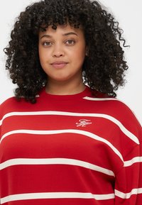 Sweatshirt rayé rouge avec des rayures horizontales blanches, présentant un col arrondi et un petit logo sur la poitrine. Texture de tissu douce.