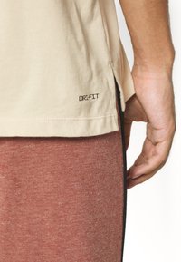 Camiseta Dri-FIT beige con un corte lateral, textura suave y un ajuste ceñido. Acompañada de pantalones ajustados de color rojo oscuro con un sutil patrón acanalado.