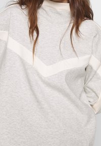 Hellgrauer Sweatshirt mit einem weißen Chevron-Muster über der Brust. Aus weichem Material gefertigt, mit einem hohen Kragen und lockerer Passform.