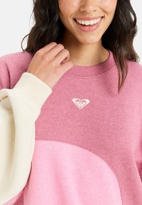 Pullover sweatshirt med gradient i pink nuancer, ribbet halsudskæring og et ærme i cremefarve. Den har et lille hjerte-logo på brystet.