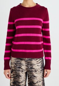 Personne portant un pull bordeaux à rayures horizontales roses et un pantalon beige avec un motif de rayures de tigre noir et gris.