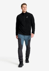 Schwarzer Fleece-Pullover mit halben Reißverschluss, kombiniert mit blauen und schwarzen Hybrid-Hosen mit strukturierten Einsätzen und verstellbaren Bündchen, getragen mit schwarzen Stiefeln.