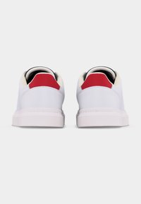 Ζευγάρι λευκών χαμηλών sneakers με κόκκινες ετικέτες στη φτέρνα και μπεζ εσωτερικά, που φαίνονται από πίσω σε απλό λευκό φόντο.