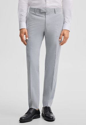 Uomo che indossa pantaloni eleganti grigio chiaro, scarpe eleganti nere e una camicia bianca con le mani rilassate lungo i fianchi.