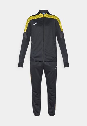 Musta urheiluharjoituspuku, jossa on keltainen kuviollinen korostus takissa. Sisältää vetoketjukiinnityksen edessä ja logon sekä takissa että housuissa.