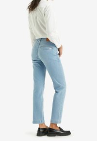 Žena ve světle modrých úzkých džínách, bílé košili s dlouhým rukávem a černých mokasínech, stojí s jednou rukou v kapse a dívá se do strany.