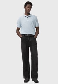 Młody mężczyzna stoi z rękami w kieszeniach, ubrany w jasnoniebieską koszulkę polo, czarny pasek, szerokie czarne jeansy i czarne buty na jednolitym tle.