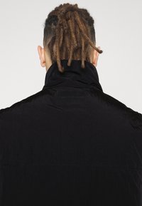 Czarna puchowa kurtka z wysokim kołnierzem i miękką, teksturowaną powierzchnią. Na kołnierzu znajduje się prostokątna etykieta z nazwą marki.