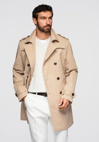 Homme barbu portant un trench-coat beige, un pull blanc et un pantalon blanc, debout avec la main gauche dans la poche, regardant vers la droite.
