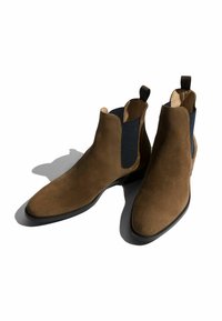 Botas Chelsea de ante marrón con paneles laterales elásticos, punta redondeada y suelas de goma negras. Incluye un tirador trasero para facilitar su uso.