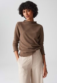 Donna con capelli ricci indossa un top lungo a maniche e texture marrone e pantaloni beige a vita alta, in piedi con una mano in tasca contro uno sfondo neutro.