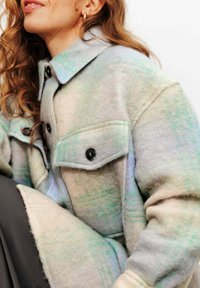 Manteau en mélange de laine gris clair avec des motifs subtils verts et bleus, doté d'un col structuré, de deux poches poitrine et de boutons noirs.