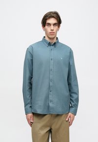 Srajca z gumbi v temni zeleni barvi, izdelana iz mešanice bombaža, z nežno teksturo. Ima ovratnik, dolge rokave in majhno logo na prsnem delu.