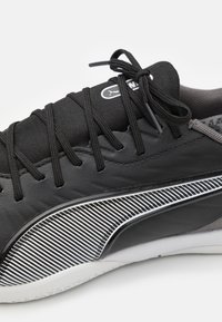 Zapatilla deportiva negra con parte superior de cuero texturizado, cordones de tela y un diseño de rayas blancas en el costado. Suela de goma con un acabado suave.