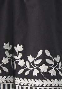 Tessuto nero con ricami floreali bianchi intricati lungo l'orlo e il lato, che combinano foglie e fiori in un motivo dettagliato.