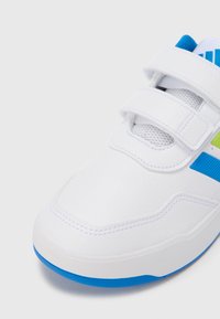 Balta sportinė avalynė su tekstūruotu viršumi, mėlynomis ir žaliomis detalėmis, Velcro užsegimais ir apvalia nosimi. Stora guminė pado dalis.