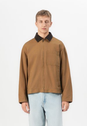 Jeune homme portant une veste zippée marron avec un col noir et un jean bleu clair, se tenant devant un fond blanc uni.