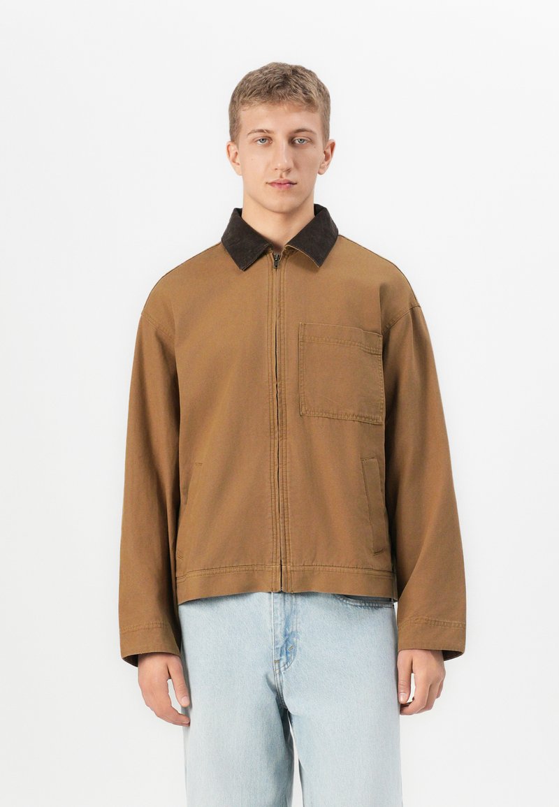 Ung mann iført en brun zip-up jakke med svart krage og lyseblå jeans, som står mot en enkel hvit bakgrunn.