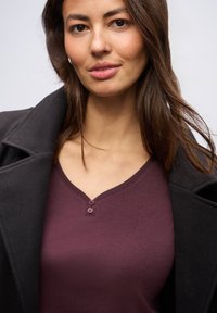 V-hals, bordeaux top med to knapper ved halsen, båret under en sort, kravejakke. Glat stofstruktur uden synlige mønstre.