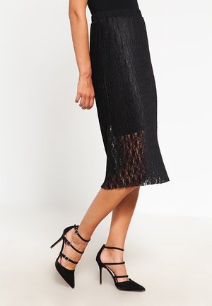 Femme portant une jupe midi noire texturée avec un ourlet en dentelle et des chaussures noires à talons hauts à bout pointu avec plusieurs brides à la cheville.
