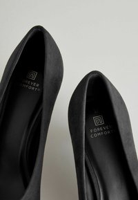 Czarne zamszowe szpilki z opływowym noskiem, z gładkim wnętrzem i wytłoczonym logo "Forever Comfort" na wkładce.