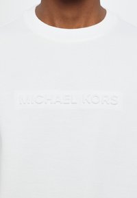 White cotton sweatshirt featuring a raised, embossed "MICHAEL KORS" logo across the chest and a classic crew neckline. Soft texture.