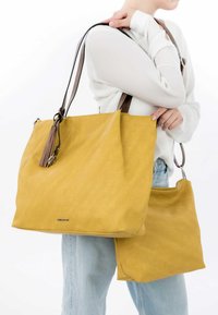 Gele canvas draagtas met bruine leren handvatten en een kwastaccent, met een kleinere bijpassende tas bevestigd. Zachte textuur, rechthoekige vorm.