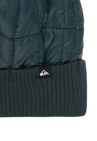 Prešívaná tmavozelená čiapka s rebrovaným tmavozeleným lemom. Obsahuje malú čiernu logovú nálepku na leme. Textúra pôsobí hladko a jemne.