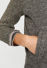 Grå texturerad tröja med gyllene trådar, med rundad halsringning och ribbade ärmslut. Ärmslutet har svarta randiga accenter.