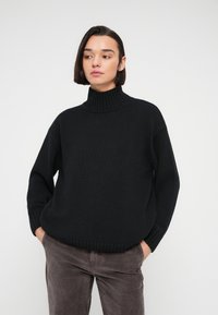 Femme portant un pull à col roulé noir et un pantalon en velours côtelé gris, les mains dans les poches, regardant légèrement sur le côté sur un fond uni.