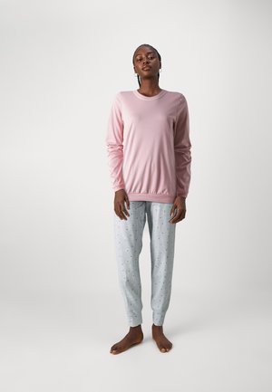 Rosa Langarmshirt aus glattem Stoff; graue Schlafanzughosen mit dezenten Mustern und Bündchen an den Knöcheln. Modell steht barfuß vor einem schlichten Hintergrund.