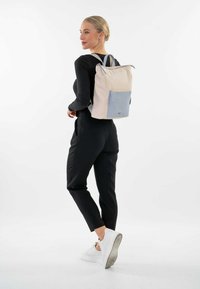 Mochila en crema con un bolsillo azul claro, hecha de tela. Cuenta con cierre de cremallera y correas ajustables. Modelo lleva puestos pantalones negros y zapatillas.