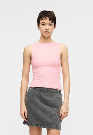 Vrouw met kort donker haar, gekleed in een mouwloze roze top en een donkergrijze denim rok, staand tegen een effen lichte achtergrond.