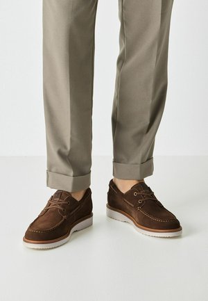 Braune Bootsschuhe aus Wildleder mit weißen Gummisohlen, versehen mit einem Schnürdesign und Nahtdetails. Kombiniert mit hochgekrempelten beigen Hosen.