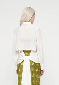 Balta trumpa bluzė su ilgomis pūslių rankovėmis, akcentuota mygtukais. Dėvima virš žalios sijono su akimis ir gėlių raštais.