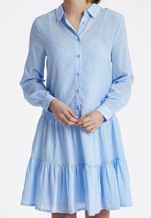 Femme portant une robe bleu clair à manches longues boutonnée, avec un motif à carreaux et un ourlet à volants superposés, les mains jointes devant.
