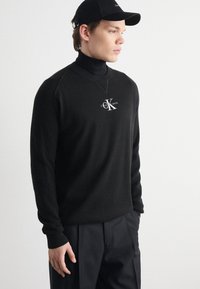 Black knit sweater with ribbed accents, high collar, and logo detail on the chest. Worn with black trousers and a matching cap.