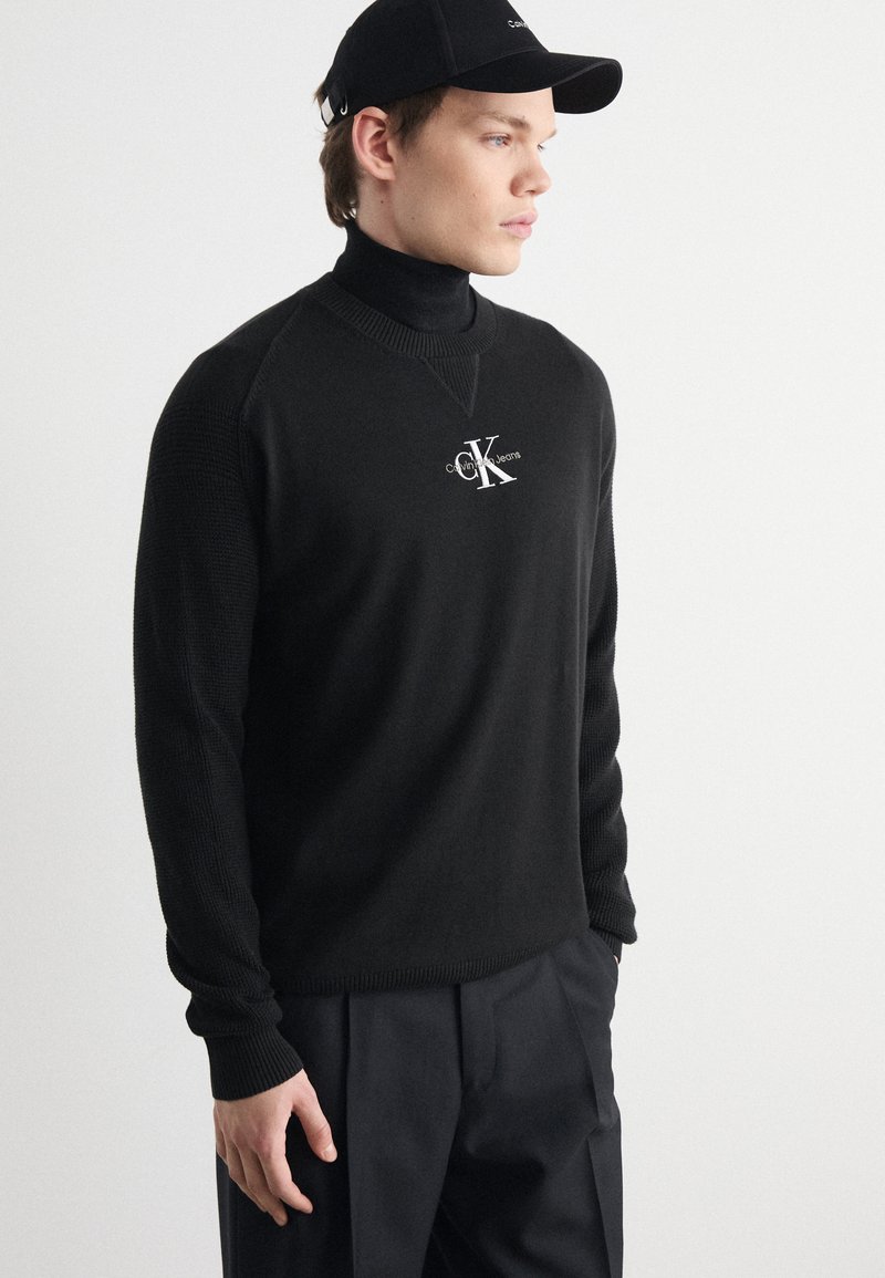 Black knit sweater with ribbed accents, high collar, and logo detail on the chest. Worn with black trousers and a matching cap.