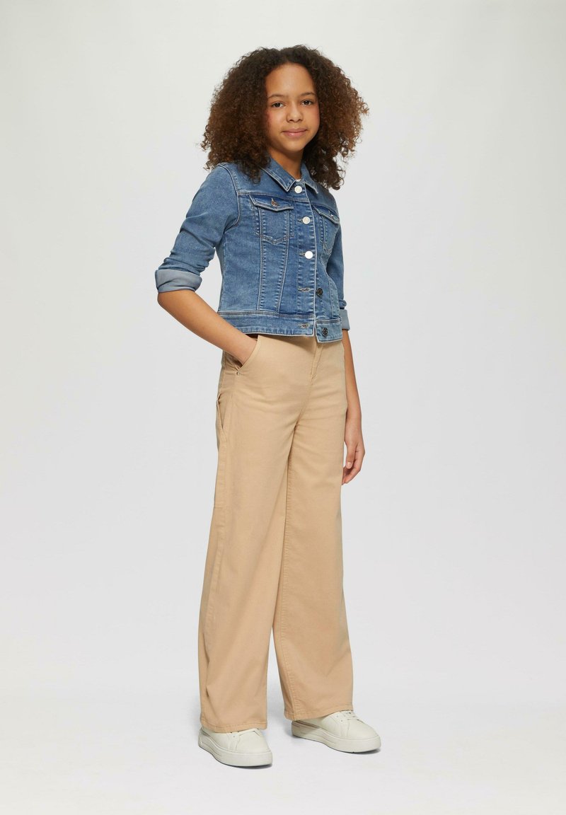 Denimjas met een getailleerd ontwerp, lichtblauwe kleur en zilveren knopen, gecombineerd met beige broek met wijdere pijpen en witte schoenen.