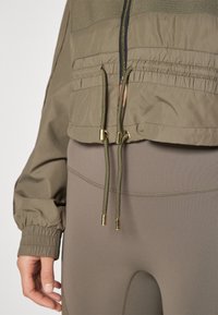 Chaqueta corta verde oliva con puños elásticos, que presenta una cremallera frontal y cordones con herrajes dorados. Combinada con unos pantalones ajustados de color marrón claro.