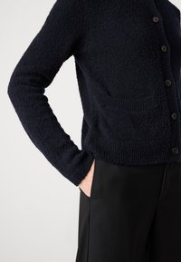 Person in einem strukturierten, dunkelblauen, geknöpften Cardigan mit Taschen und schwarzen Hosen, eine Hand in der Tasche, vor einem schlichten Hintergrund.