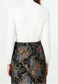 Kvinna i en vit långärmad blus instoppad i en högmidjad svart kjol med ett brunt och blått blommönster, sedd bakifrån.