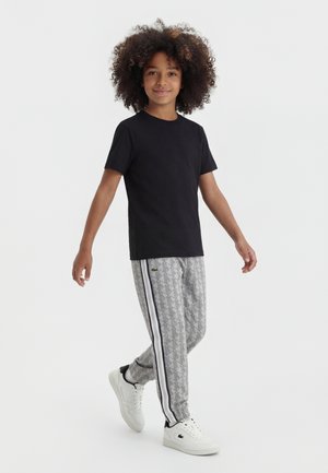 Enfant aux cheveux bouclés marchant, portant un t-shirt noir, un pantalon gris à motifs avec des bandes latérales noires et blanches, et des baskets blanches avec un détail noir.