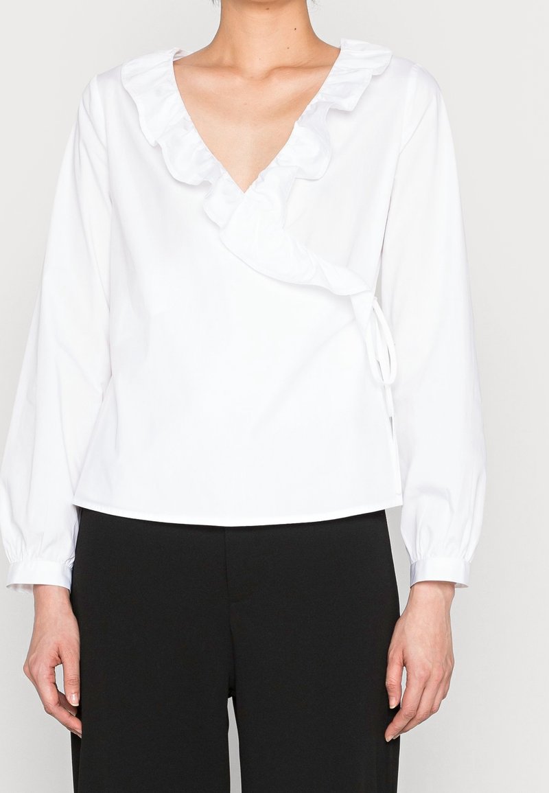 Blusa bianca a maniche lunghe con scollo a V e collo arricciato. Presenta un design avvolgente con legature laterali e polsini con bottoni. Tessuto dalla texture liscia.