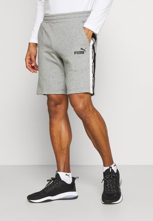 Man draagt grijze Puma-short met merkgestreepte zijkanten, witte Puma-sokken en zwarte Puma-sneakers, staand tegen een witte muur.