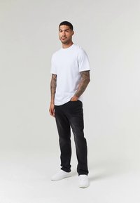 T-shirt branca de manga curta com decote redondo, combinada com jeans pretos e ténis brancos. O modelo tem tatuagens visíveis nos braços.