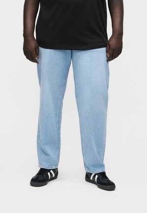 Stovintis asmuo dėvi šviesiai mėlynas laisvo silueto džinsus, juodus sportbačius su baltais dryžiais ir juodą marškinėlį, matomas nuo krūtinės vidurio žemyn.