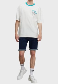 Hombre con una camiseta gráfica blanca de cuello aguamarina, pantalones cortos de mezclilla oscuros y doblados, calcetines blancos con rayas negras y zapatillas blancas.